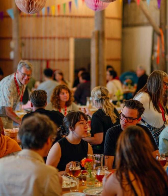 Gæster sidder ved langborde i en rustik lade hos Herslev Bryghus, omgivet af farverige papirdekorationer og hyggelig belysning.