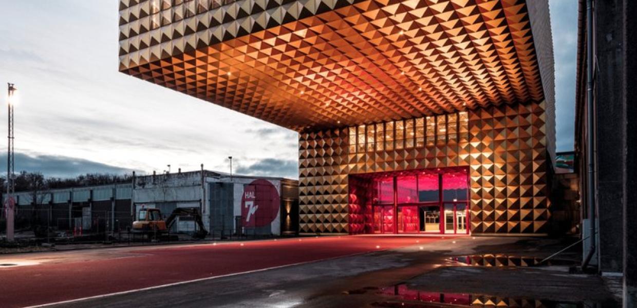 Gylden, kubeformet facade på Ragnarock med markant udhæng og rødlyst indgang i aftenskumring.