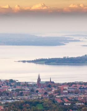 Udsigt over Roskilde med Roskilde Domkirke i forgrunden og Roskilde Fjord omgivet af øer og landskab i baggrunden under en himmel med skyer i aftensol.