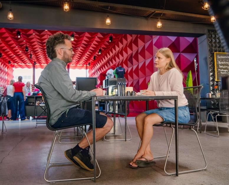 To personer sidder ved et cafébord i Ragnarocks karakteristiske røde, geometriske interiør, mens andre gæster bevæger sig i baggrunden.