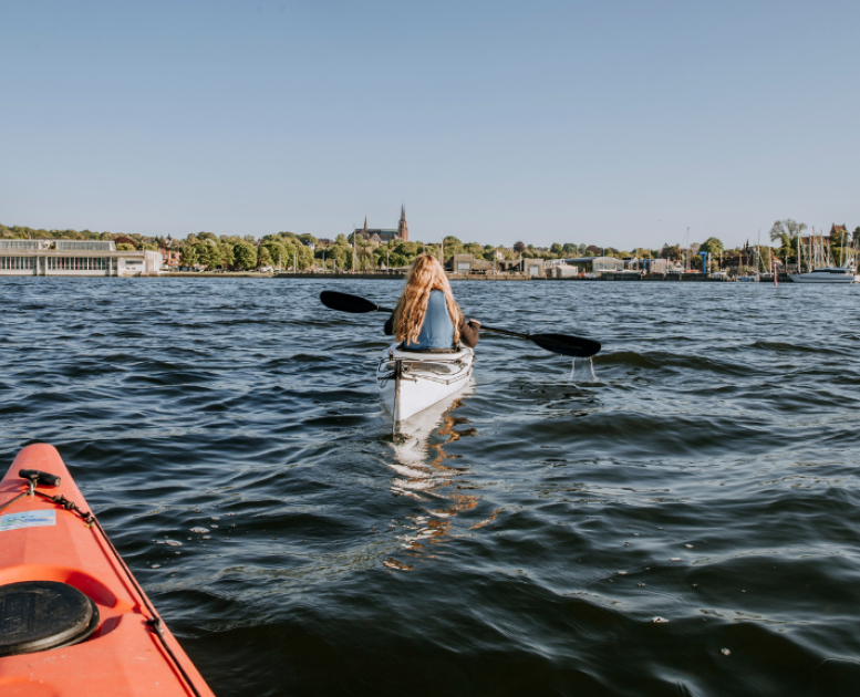 Person sejler kajak på Roskilde Fjord med udsigt til byen og havnen i baggrunden.